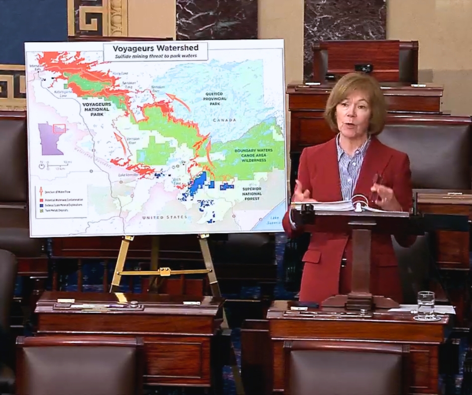 A female stands at a podium giving a speech with a poster board in the background.