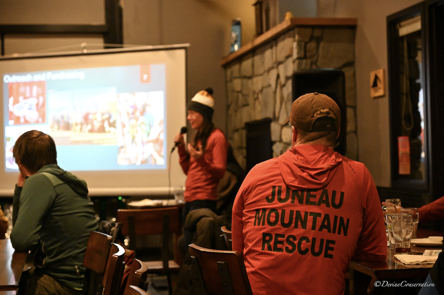 A bald man wearing a shirt that says Juneau Mountain Rescue is sitting in a large room in front of a screen and a presenter. 