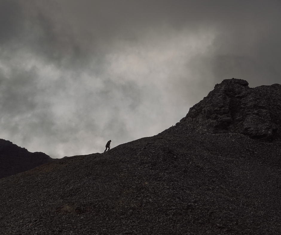 Man walks up mountain. Grey skies filled with clouds.