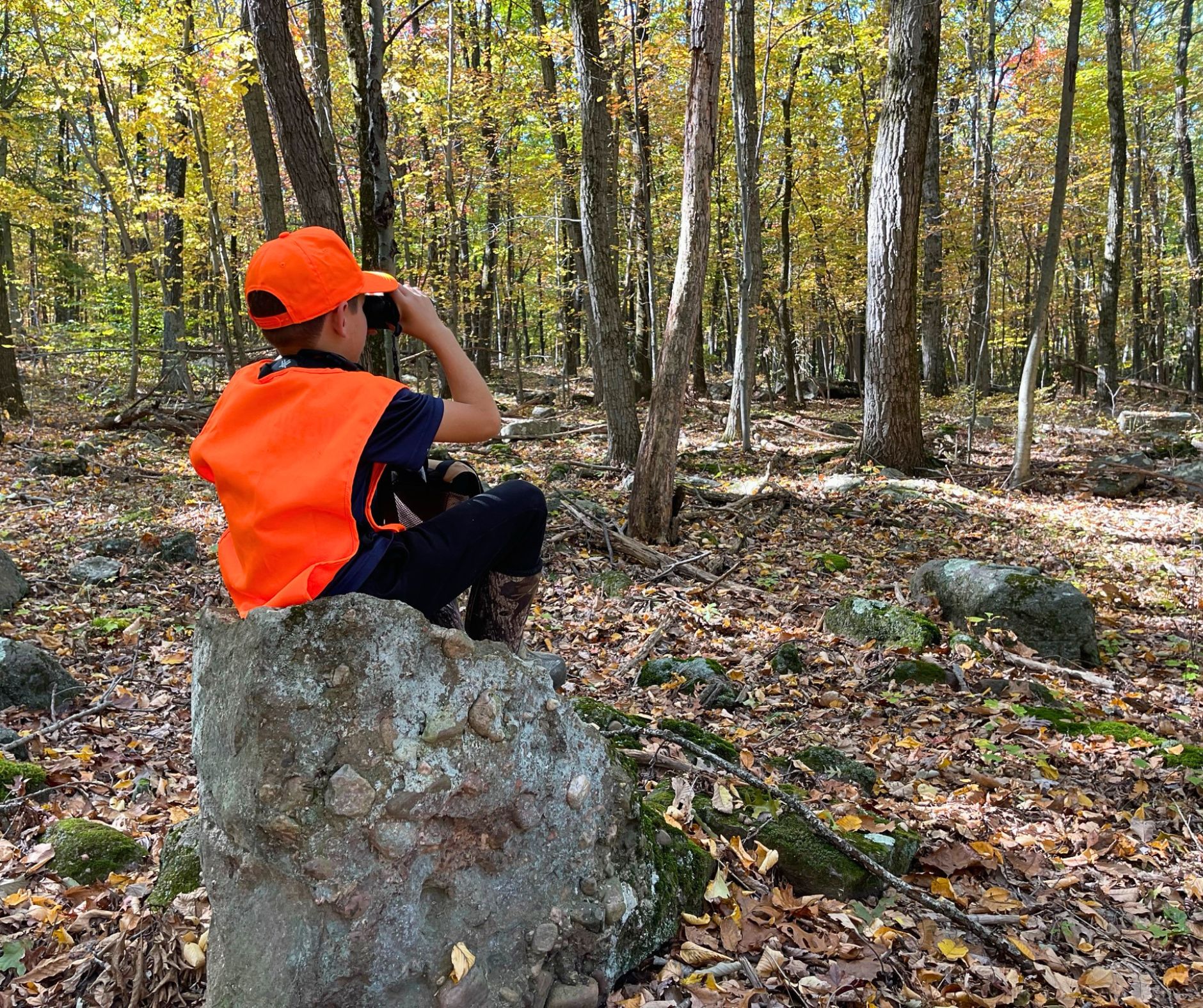 A young boy sits atop a rock while wearing a blaze orange vest and looking through binoculars.